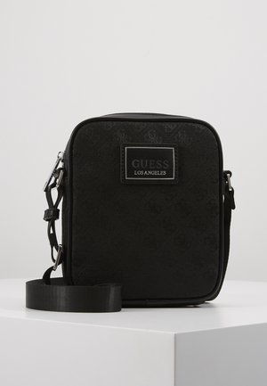 Czarna torebka na ramię w kształcie kwadratu z regulowanym paskiem i naszywką z logo "GUESS Los Angeles" na przednim panelu, przedstawiona na białym tle.