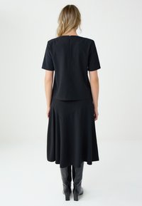 Kobieta stojąca tyłem, ubrana w czarną bluzkę z krótkim rękawem, czarną spódnicę midi i czarne kozaki do kolan na gładkim tle.