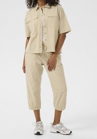 Chemise rayée beige à manches courtes avec deux poches poitrine, associée à un pantalon court assorti doté d'une taille élastique. Portée avec des baskets.