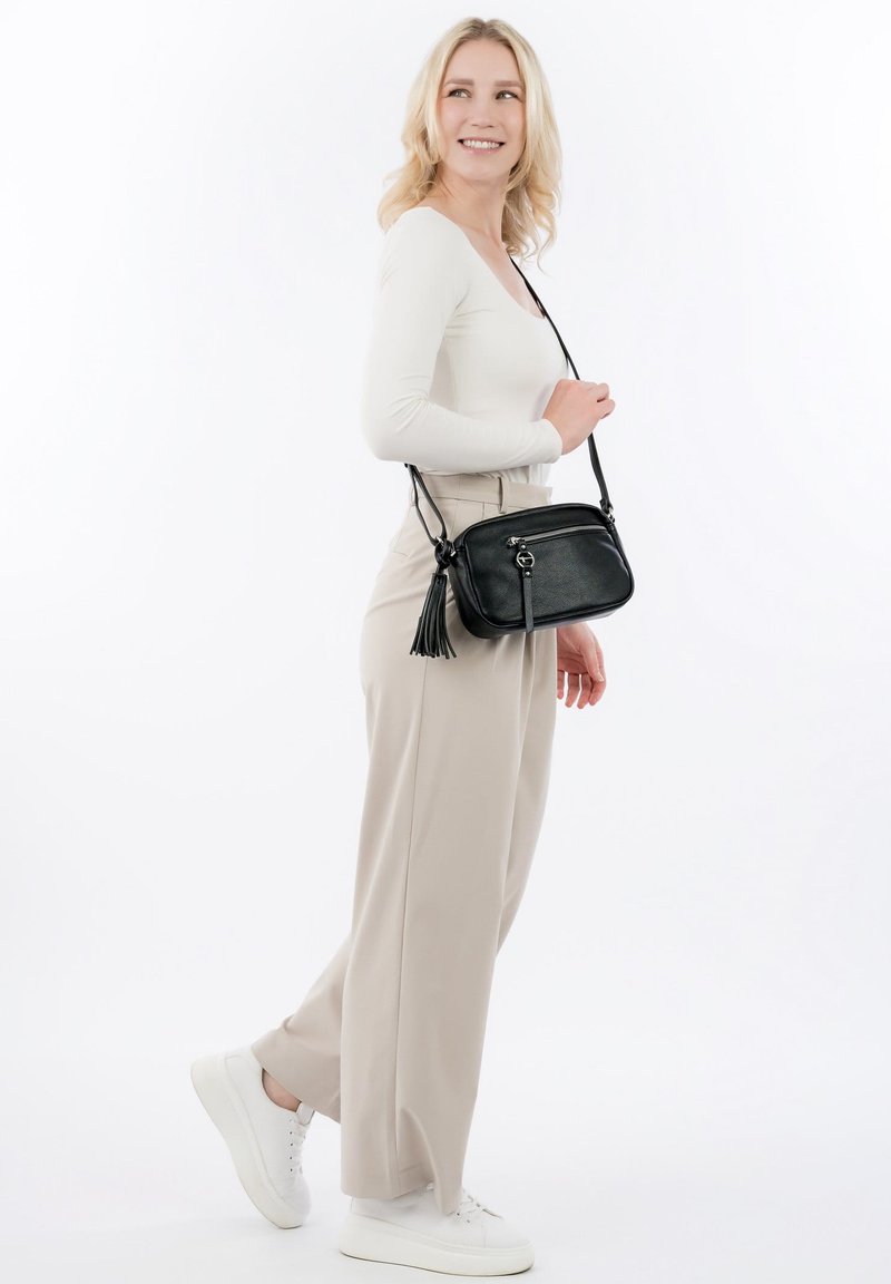 Smilende kvinde iført hvid langærmet top, beige bukser med vide ben, hvide sneakers, bærende på en sort crossbody-taske med kvast mod hvid baggrund.