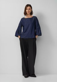 Dunkelblaues Denim-Oberteil mit weitenÄrmeln und gerafften Akzenten; kombiniert mit hoch taillierten schwarzen Hosen und spitzen schwarzen Ballerinas.