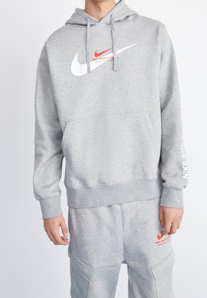 Person iført en lysegrå Nike-hættetrøje og matchende joggingbukser med hvide og røde Nike-logoer og tekst.