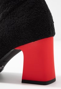 Zapato negro texturizado con un tacón bloque rojo. El tacón presenta una superficie lisa que contrasta con la tela esponjosa del zapato.