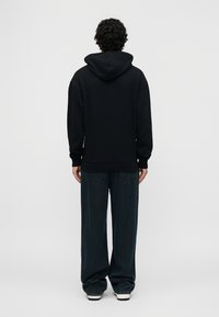 Άτομο με σγουρά μαλλιά στέκεται με την πλάτη προς τον θεατή, φοράει μαύρη φούτερ με κουκούλα, φαρδιά σκούρα τζιν και λευκά αθλητικά παπούτσια, μπροστά σε απλό ανοιχτόχρωμο φόντο.