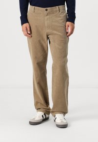 Beige Cordhosen mit geradem Schnitt, mit einem Knopfverschluss und seitlichen Taschen, getragen mit weißen Sneakers und einem dunkelblauen Langarmshirt.
