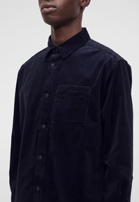 Homme portant une chemise en velours côtelé bleu marine foncé à boutons avec col et poche poitrine sur un fond clair uni.