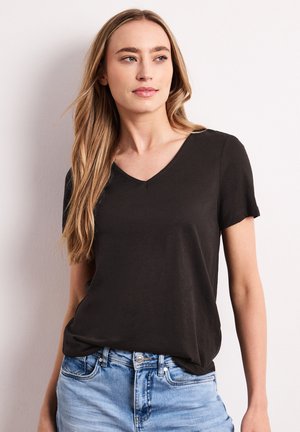 Jeune femme aux longs cheveux blonds portant un T-shirt noir à col en V et un jean bleu clair, regardant légèrement sur le côté sur un fond uni.