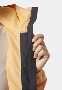 Orange jackas insida med ett nätfoder och en kontrasterande svart kant. En hand håller jackans kant, vilket visar tygtexturen.