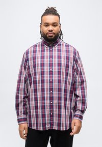 Άνδρας με ντραντλοκς και γένια, φοράει κουμπωτό καρό πουκάμισο σε κόκκινο, λευκό και μπλε χρώμα, στέκεται μπροστά σε απλό φωτεινό φόντο.