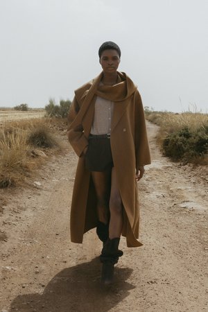 Cappotto di lana lungo e marrone con un colletto oversize, abbinato a un top grigio chiaro e pantaloni corti scuri. Indossato con stivali neri spessi alla caviglia su un sentiero di terra.
