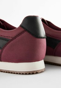 Batik sneakers merah marun dengan aksen kulit hitam, tekstur halus, jahitan kontras, dan sol karet putih yang menampilkan pola zigzag.