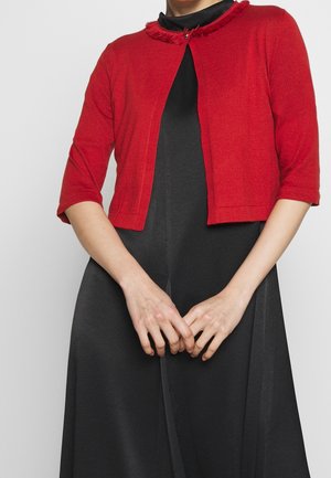 Kobieta w czerwonym krótkim kardiganie z rękawami do połowy łokcia, na czarnej sukience z wysokim kołnierzem, z rękami złożonymi na przodzie.