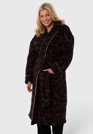 Mujer sonriente que lleva una bata de forro polar con estampado de leopardo de color marrón oscuro, ribete rosa y un cinturón en la cintura, de pie con una mano en el bolsillo.