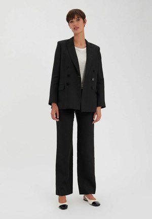 Blazer noir à double boutonnage avec des revers larges, associé à un pantalon large assorti et des ballerines bicolores. Le tissu semble lisse.