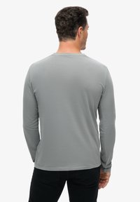 Man met kort krullend haar, gekleed in een lichtgrijze lange mouwen shirt en zwarte broek, met zijn rug naar de camera tegen een eenvoudige witte achtergrond.