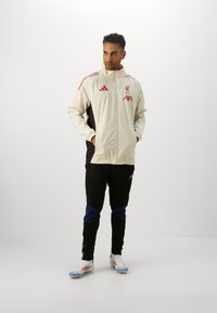 Adidas jakke i cremefarvet med sorte sider, prydet med Liverpool-logo og røde detaljer. Sorte joggers med blå detaljer. Hvide fodboldstøvler med farvede detaljer.
