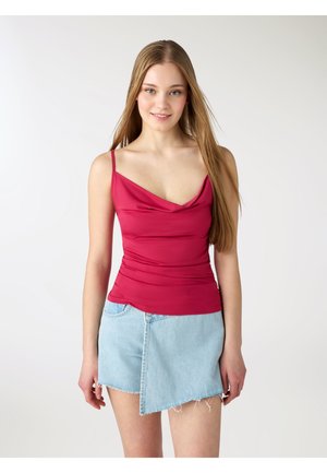 Giovane donna con capelli lunghi che indossa un top rosso senza maniche e una gonna di jeans azzurra asimmetrica, in piedi davanti a uno sfondo neutro.