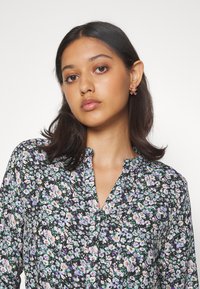 Svart blommig blus med lila, vita och gröna mönster. Har en knappslå framtill och en mandarin-krage. Mjuk tygtextur.