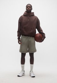 Άνδρας που φοράει καφέ φούτερ με κουκούλα και γκρι σορτς μπάσκετ κρατάει μια μπάλα μπάσκετ με το λογότυπο της Jordan, στέκεται σε λευκό φόντο.