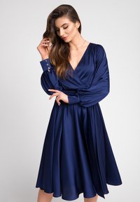 Niebieska satynowa sukienka z dekoltem w kształcie litery V, długimi bufiastymi rękawami i guzikami przy mankietach. Rozkloszowana spódnica sięga poniżej kolan.