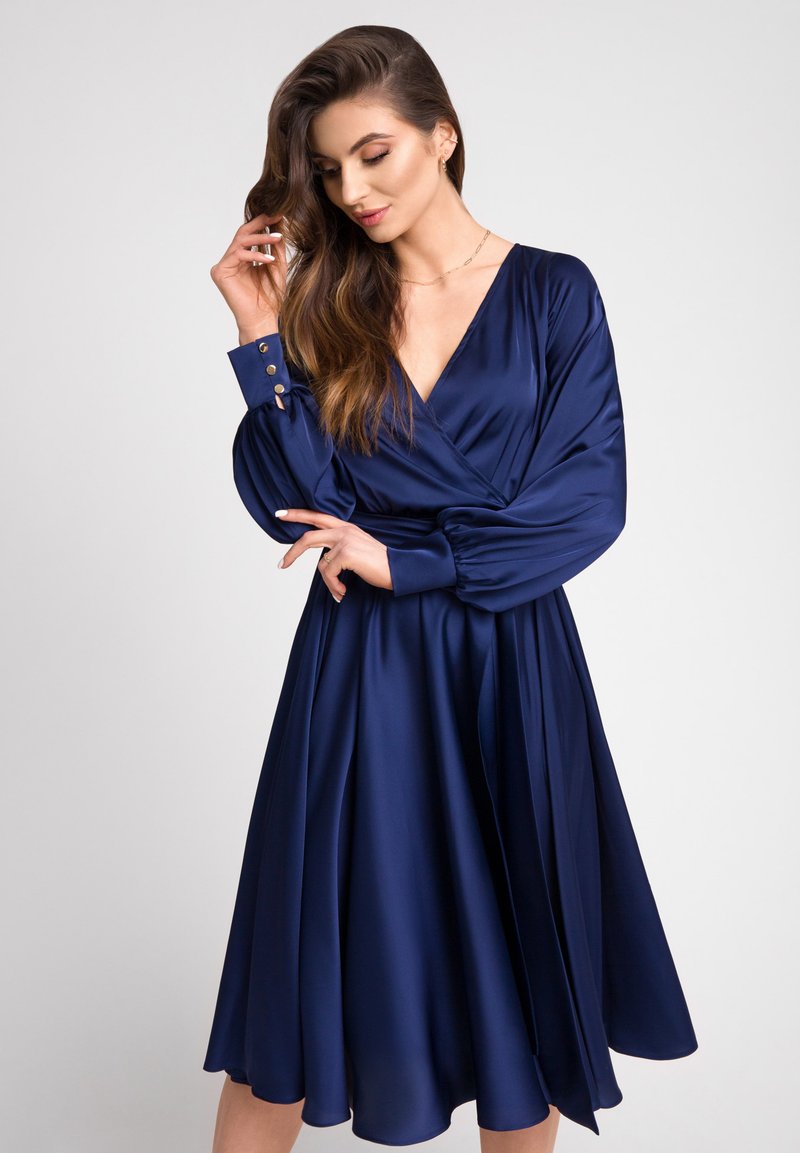 Niebieska satynowa sukienka z dekoltem w kształcie litery V, długimi bufiastymi rękawami i guzikami przy mankietach. Rozkloszowana spódnica sięga poniżej kolan.