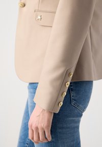 Beige sako se zlatými knoflíky a detailem kapsy, kombinované se světle modrými džíny. Tkanina vypadá hladce s vyztuženými ramenními částmi.
