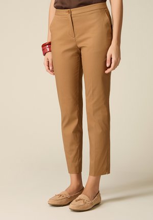 Donna che indossa pantaloni slim fit color cammello, mocassini beige e bracciali rossi al polso sinistro, in piedi su sfondo neutro.