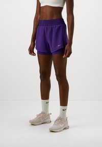 Athletisk person iført lagdelte lila shorts, hvit crop-top, hvite Nike-strømper og beige Nike løpesko, stående på en ensfarget bakgrunn.