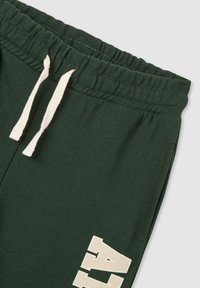 Zielone bawełniane spodnie dresowe z elastycznym pasem, kremowym sznurkiem i wyraźnym białym napisem na lewej nogawce. Miękka faktura, casualowy design.