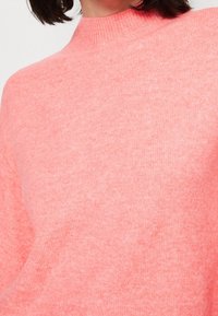 Růžový pletený svetr s vysokým límcem, vyrobený z měkké, texturované látky a volným, pohodlným střihem. Bez viditelných vzorů nebo ozdob.