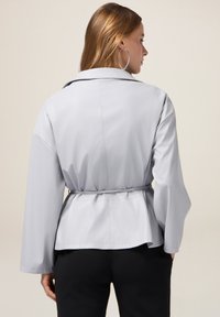Giacca grigia chiara con cintura, dal taglio morbido, maniche ampie e una texture liscia. Presenta un colletto classico e arricciature in vita.