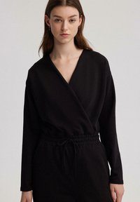 Mono negro ajustado con mangas largas, cintura elástica y detalle con cordón. Tejido suave con una textura lisa. Diseño simple y elegante.