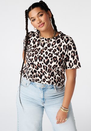 Vrouw met lange vlechten draagt een shirt met luipaardprint, lichtblauwe jeans, gouden sieraden en glimlacht tegen een effen lichte achtergrond.