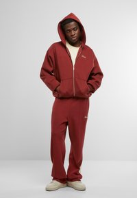 Felpa con zip di un rosso scuro con tasca frontale, abbinata a pantaloni jogger coordinati. Presenta un'accademia crema sui pantaloni e un logo ricamato su entrambi i capi.