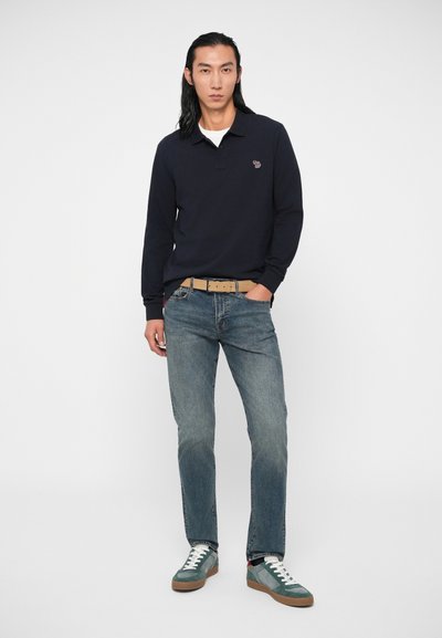 Polo de manga longa azul-marinho com gola, apresentando um pequeno logotipo. Combinado com jeans azul-claro e ténis verdes com detalhes brancos.