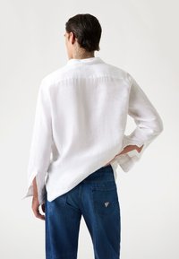 Camisa blanca de lino con mangas largas, aberturas laterales y corte relajado, combinada con jeans azul oscuro que cuentan con bolsillos traseros y detalle de logo.