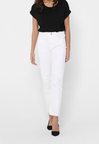 Bílé džíny s vysokým pasem, rovnými nohavicemi a roztrhaným lemem, sladěné s černým tričkem s krátkým rukávem a černými podpatky.