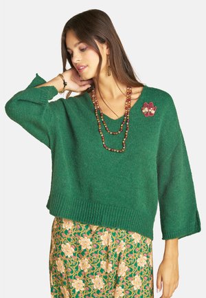 Junge Frau trägt einen grünen Pullover mit Blumenrock, Perlenkette und Ohrringen und berührt mit einer Hand ihren Hals.