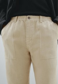 Pantalons en lin beiges avec une taille élastique, fermeture à bouton unique et deux poches avant. Tissu texturé à finition naturelle.