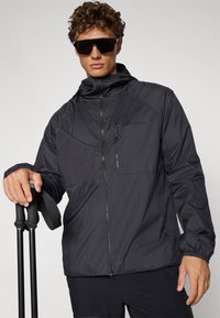 Moški v črni vetrovki in temnih sončnih očalih, ki drži črne pohodniške palice na enobarvnem ozadju.