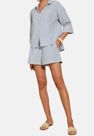 Kobieta w koszuli na guziki z pionowymi jasnoniebieskimi i białymi paskami oraz dopasowanych szortach, w beżowych sandałach wsuwanych i bransoletkach.