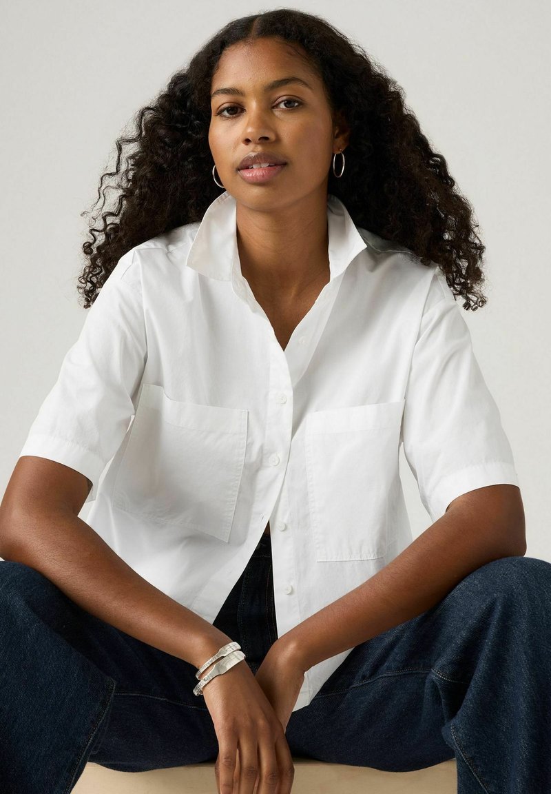 Camicia bianca a maniche corte con chiusura a bottoni, due tasche sul petto e colletto strutturato. Indossata con jeans scuri, che mostrano una texture liscia.