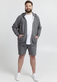 Sudadera gris con cremallera, cordones y bolsillos frontales, llevada sobre una camiseta blanca y pantalones cortos grises a juego. Los zapatos son blancos.