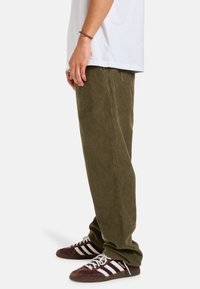 Olive green corduroy pants with a relaxed fit, side pockets, and textured fabric; paired with striped brown and white sneakers.