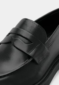 Chaussure mocassin en cuir noir à finition lisse, avec des accents décoratifs cousus et une bande surélevée traversant l'avant. Semelle plate.