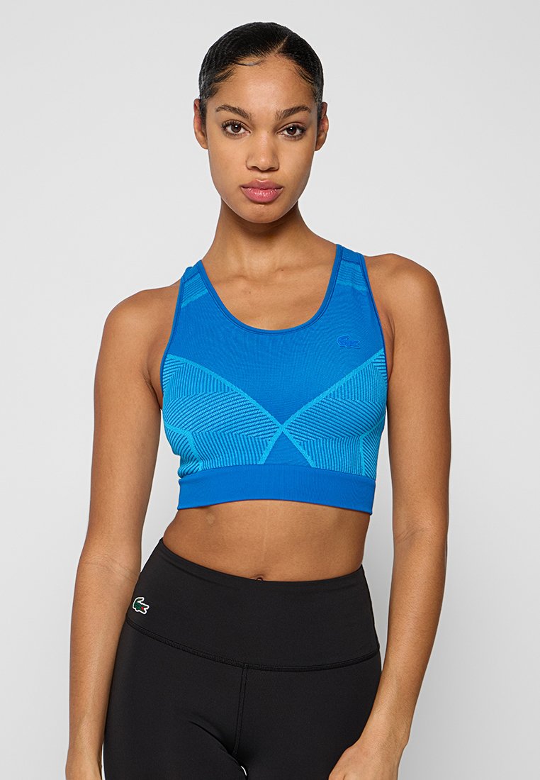 Mujer con el cabello recogido, que lleva puesta una camiseta deportiva azul con un estampado y leggings negros de cintura alta con logotipos de Lacoste en ambos.