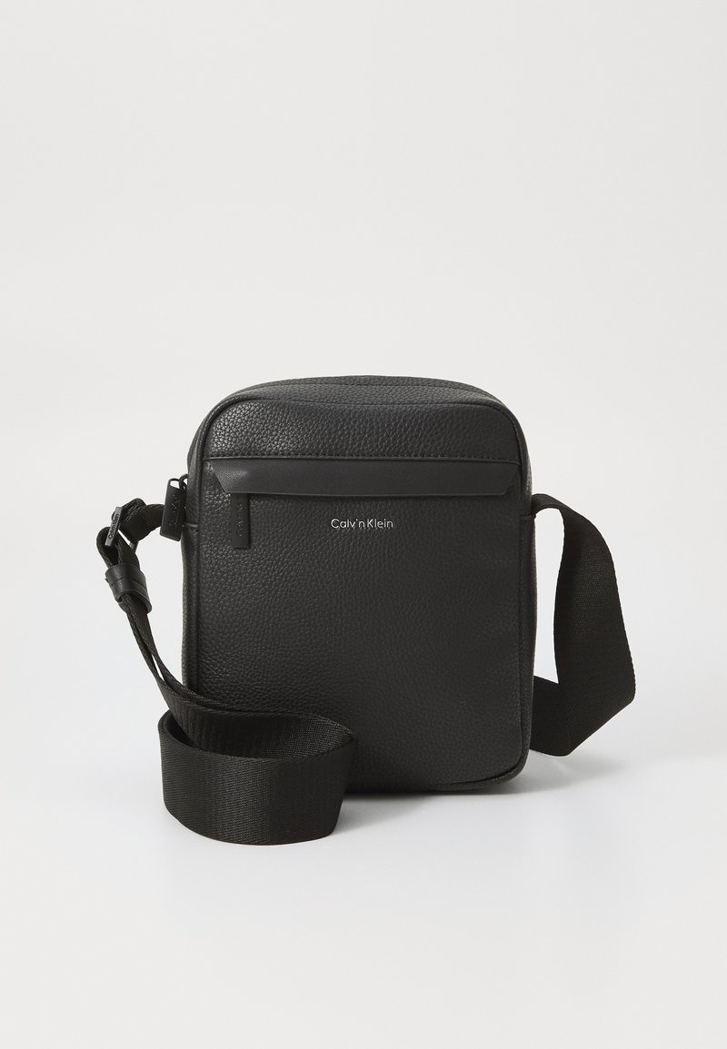 Black leather crossbody bag with a textured finish, featuring a front zipper pocket and an adjustable fabric strap. Brand logo visible.