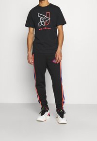 Muž v černém tričku Air Jordan s grafickým logem, v černých teplákách s oranžovými a modrými pruhy, a v bílých teniskách Air Jordan s černými tkaničkami.