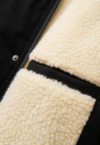 Interior de chaqueta negra con cremallera que presenta un suave y esponjoso forro de lana crema con una superficie texturizada y un panel de tela negro en contraste.