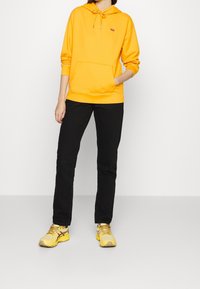 Sudadera amarilla con capucha y un bolsillo frontal, combinada con pantalones negros y zapatillas deportivas amarillas. Diseño sencillo, sin patrones visibles.